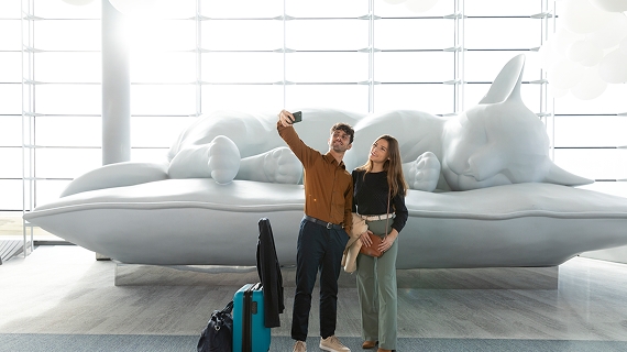 Homme et femme avec leur valise prenant un selfie devant la statue géante d'un chat dans le terminal 2E